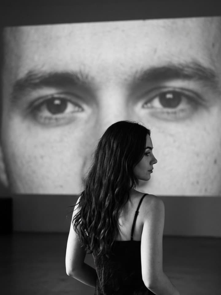 Schwarz-Weiß-Foto einer dunkelhaarigen Frau im schwarzen Slipkleid, mit dem Rücken zur Kamera, vor einer riesigen Projektion der Augen eines Mannes