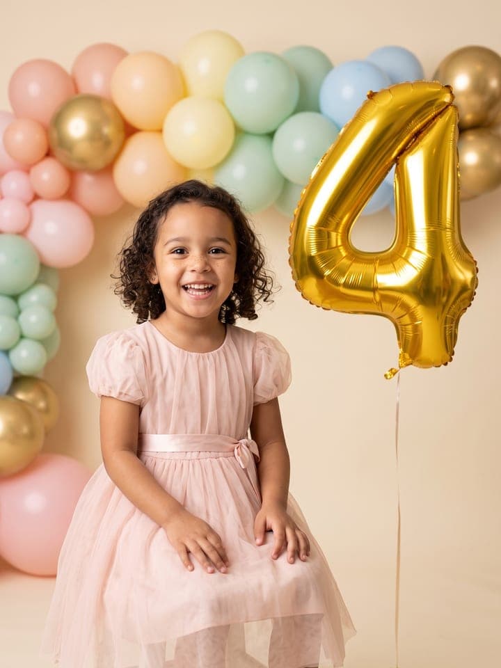 Kleines Mädchen mit dunklen Locken im zartrosa Tüllkleid lacht mit den Händen im Schoß in einem Ballonstudio in Pastell, neben ihr schwebt ein riesiger goldener Folienballon in Form einer 4