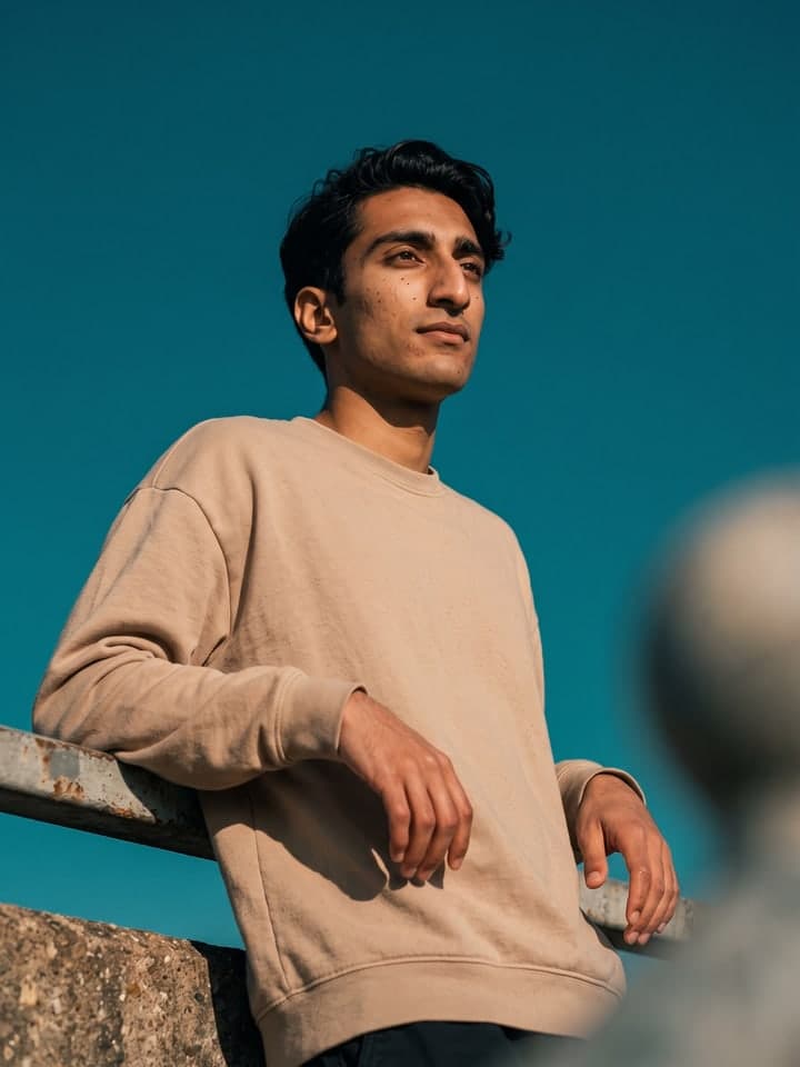 Mann mit welligen schwarzen Haaren in übergroßem beigem Sweatshirt lehnt sich an ein rostiges Geländer aus Beton und Metall, von unten vor einem durchgehend petrolfarbenen Himmel fotografiert, mit Blick nach rechts im warmen Sonnenlicht