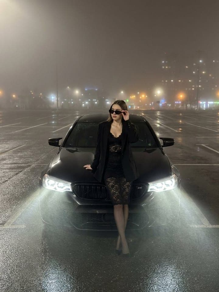 Woman in black leaning on the hood of a black BMW M5 in night fog, headlights beaming