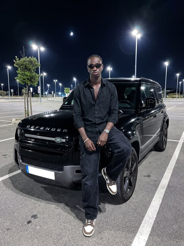 Junger Mann mit kurzen Braids in schwarzem Hemd mit hochgekrempelten Ärmeln, weiter schwarzer Cargo-Jeans und braun-weißen Sneakern sitzt nachts halb auf dem vorderen Kotflügel eines schwarzen Land Rover Defender auf einem Parkplatz, Sonnenbrille auf, dahinter Straßenlaternen und eine Mondsichel