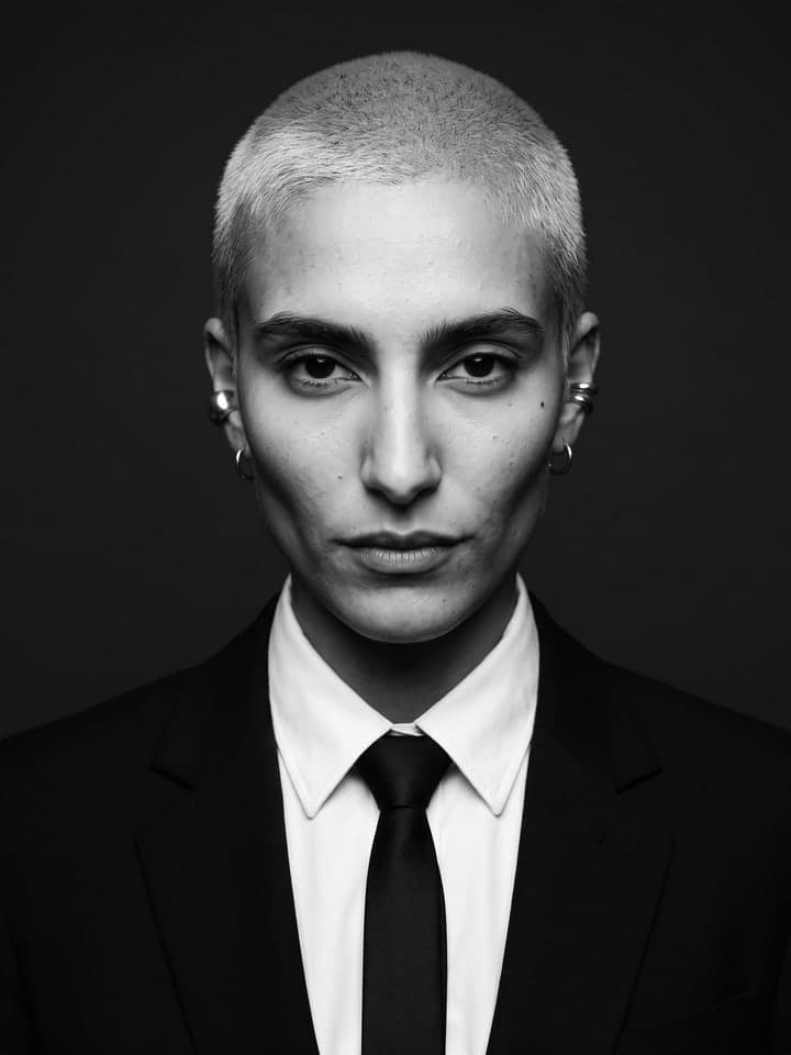 Schwarz-Weiß-Studioporträt einer Frau mit blondiertem Buzz Cut und silbernen Ear Cuffs in schwarzem Anzugsakko, weißem Hemd mit Kragen und schmaler schwarzer Krawatte, ernster direkter Blick in die Kamera, dunkler gleichmäßiger Hintergrund