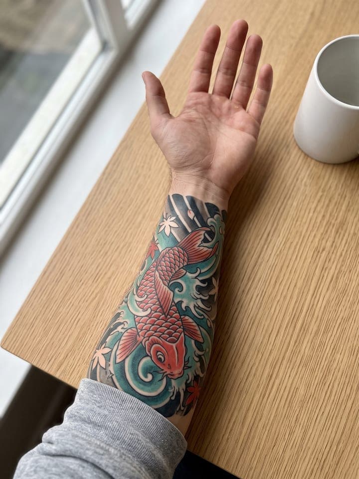 Die Innenseite des Unterarms eines Mannes, mit der Handfläche nach oben auf einem Eichentisch am Fenster, mit einem verheilten Tattoo im japanischen Stil: ein roter Koi in blaugrünen Wellen und kleine Ahornblätter, die sich um den Arm legen