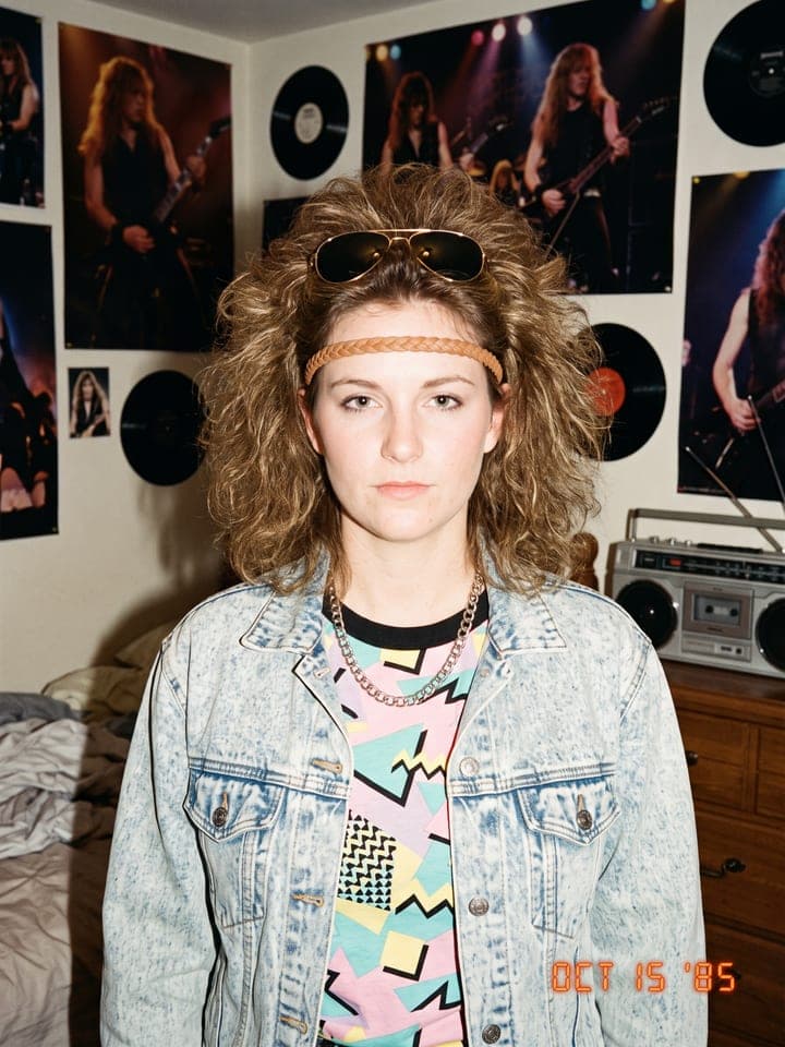 Junge Frau mit großer 80er-Dauerwelle, geflochtenem Haarband, Pilotenbrille und Acid-Wash-Jeansjacke in einem Zimmer voller Poster, Datumsstempel OCT 15 '85