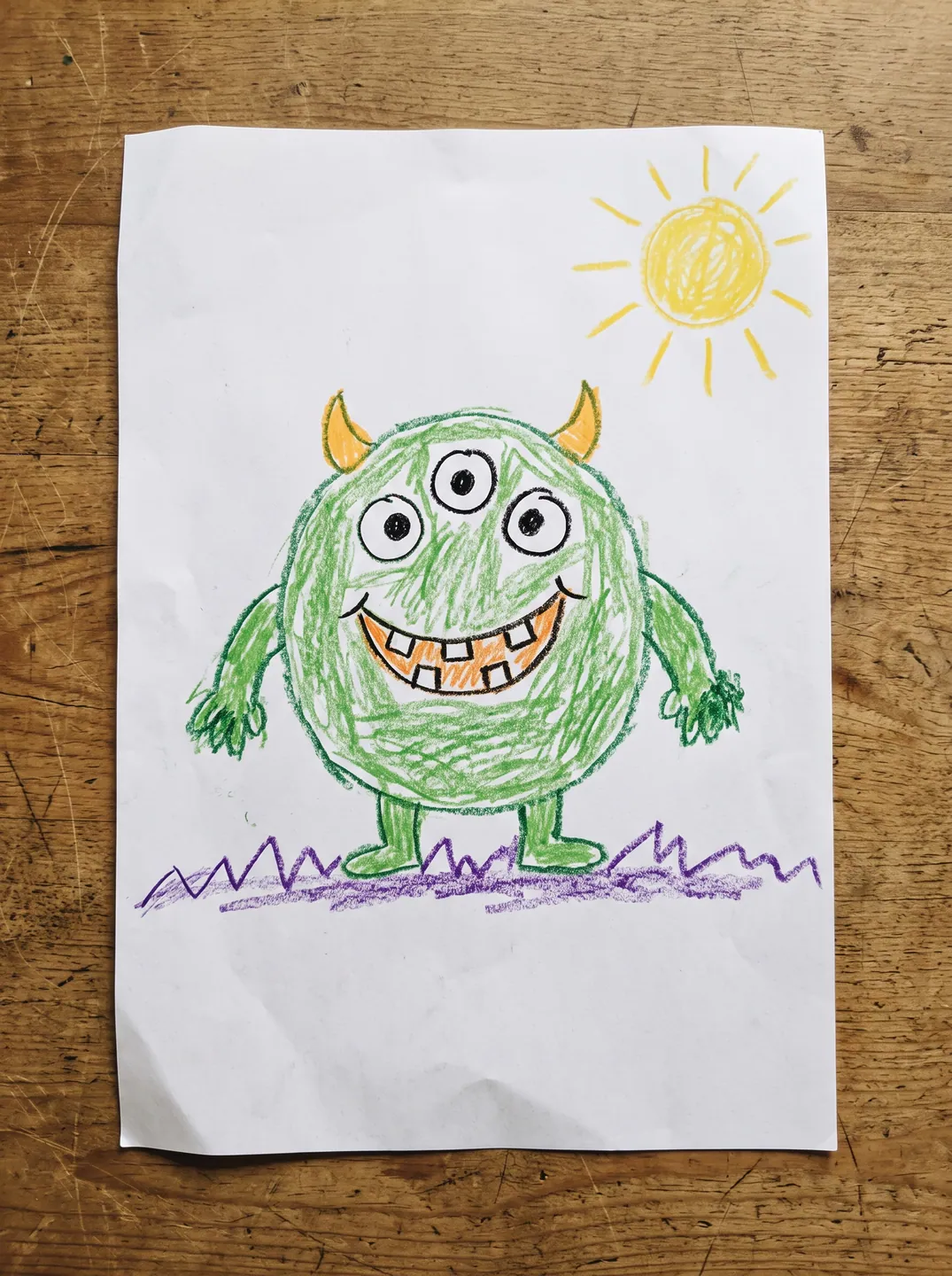 Crayon drawing of a green three-eyed monster with orange horns on a sheet on a wooden table, used as the input