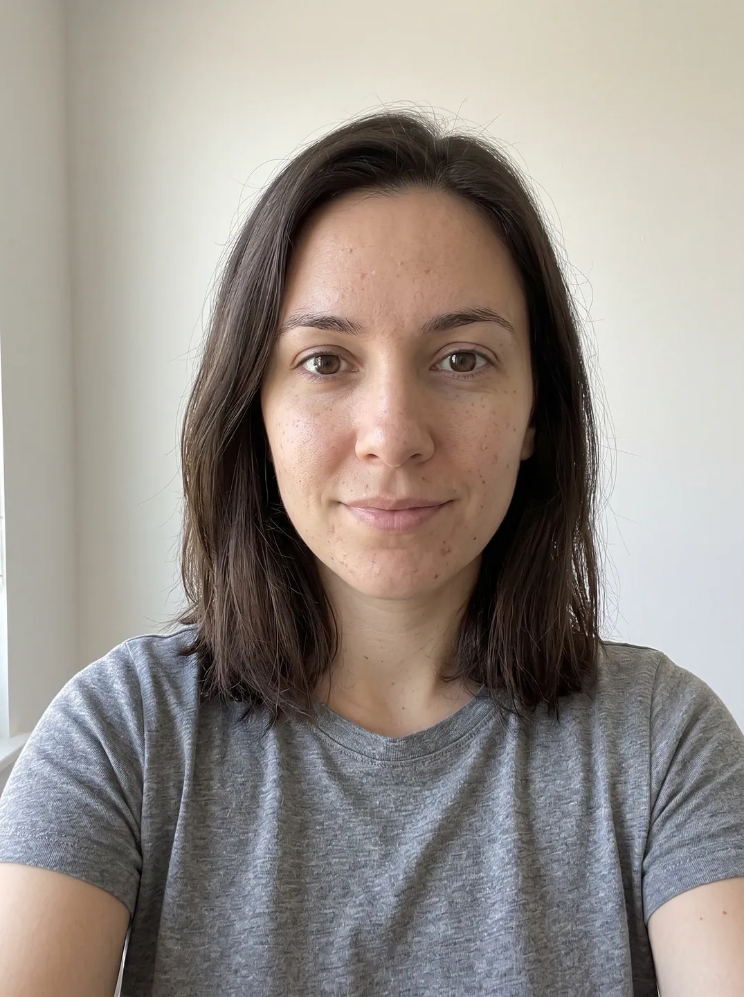Selfie of a woman with shoulder-length dark brown hair in a gray T-shirt against a white wall, used as the input